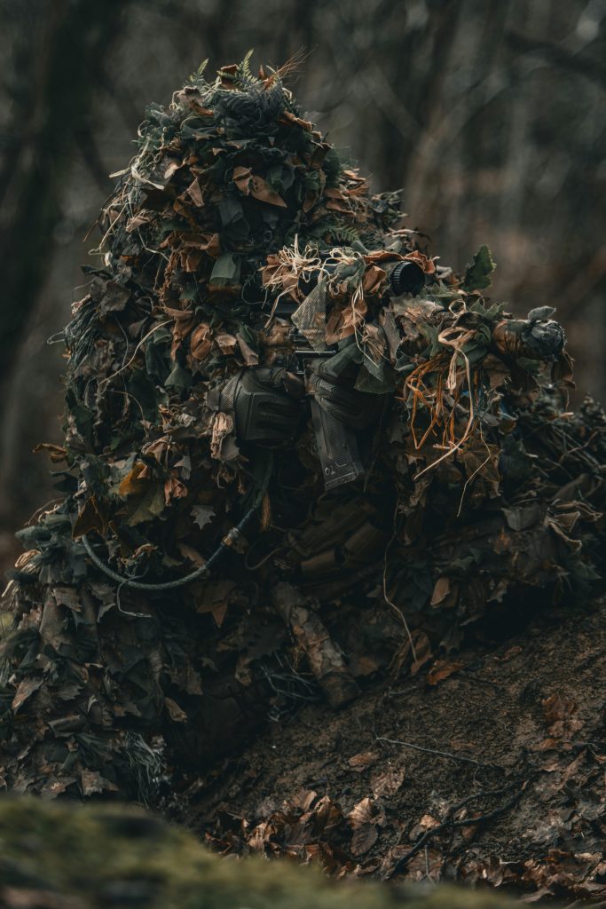 pexels-photo-17725277-17725277 A sniper in camouflage lying on the ground covered in leaves in a forest setting, aiming intently.