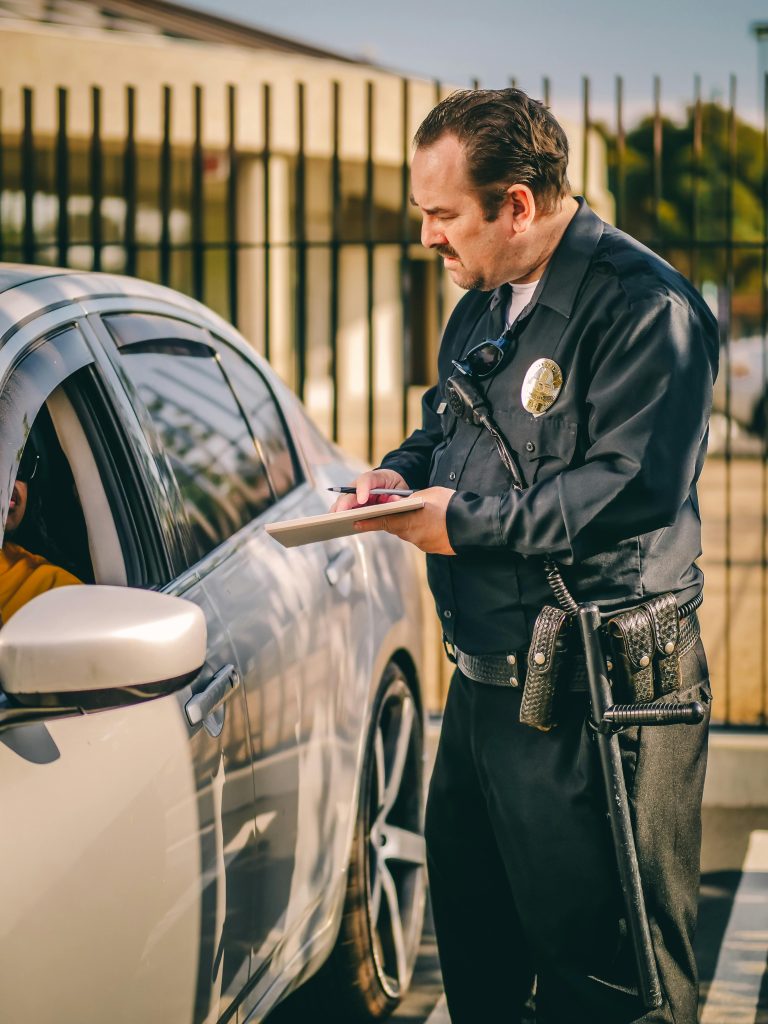 pexels-photo-7715236-7715236 A police officer writes a speeding ticket for a driver in a silver car on a sunny day.