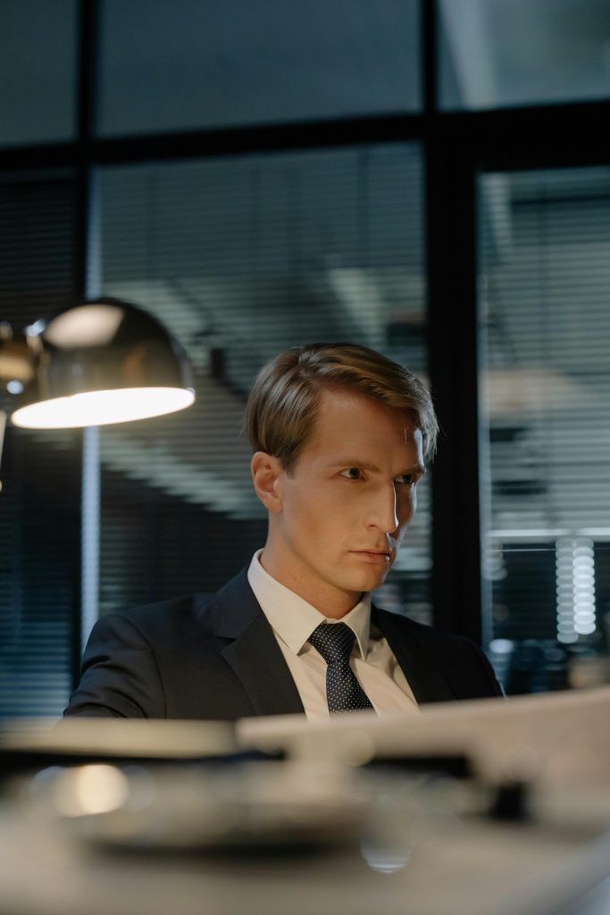 pexels-photo-8382287-8382287 Serious detective in a suit examining documents under a desk lamp at night in a modern office.